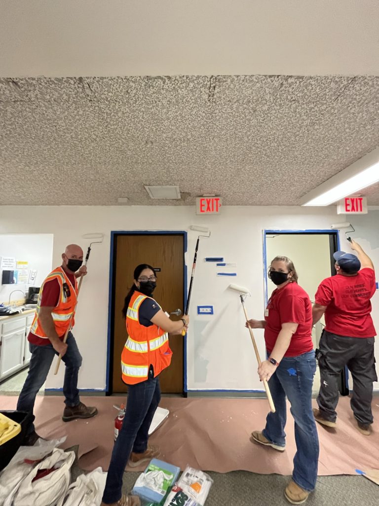 Four people are painting the walls of an indoor space. They are holding paint rollers on long handles, working together on a renovation project. Two of the participants are wearing orange reflective vests, and all are wearing masks for safety. The scene is part of a community service team building activity, with individuals collaborating to repaint the space.