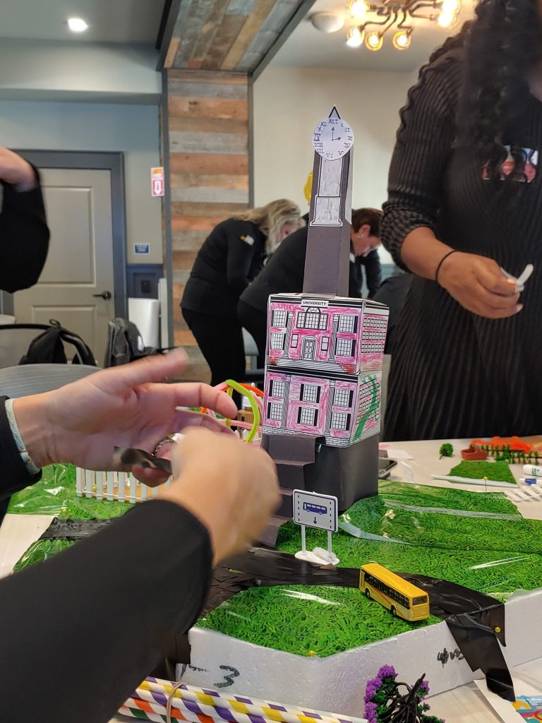 Close-up of a creative tower structure with a clock and decorated buildings in a City Build team building event, surrounded by miniature roads and greenery.