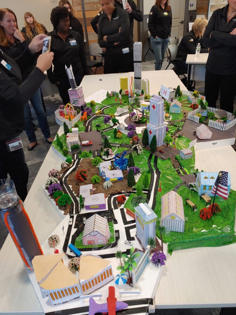 Participants at a City Build team building event admire and photograph an intricately designed miniature cityscape featuring roads, buildings, trees, and a central clock tower.