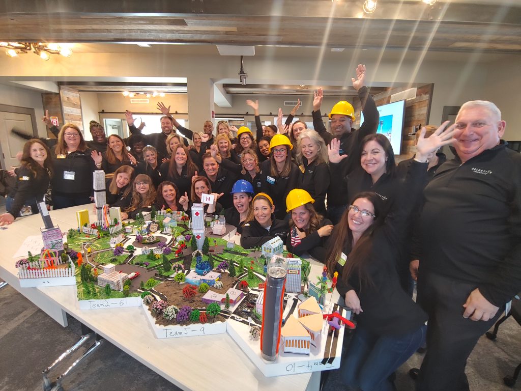 A large group of participants celebrates their success at a City Build team building event, posing around a detailed miniature city model while wearing hard hats and smiling enthusiastically.