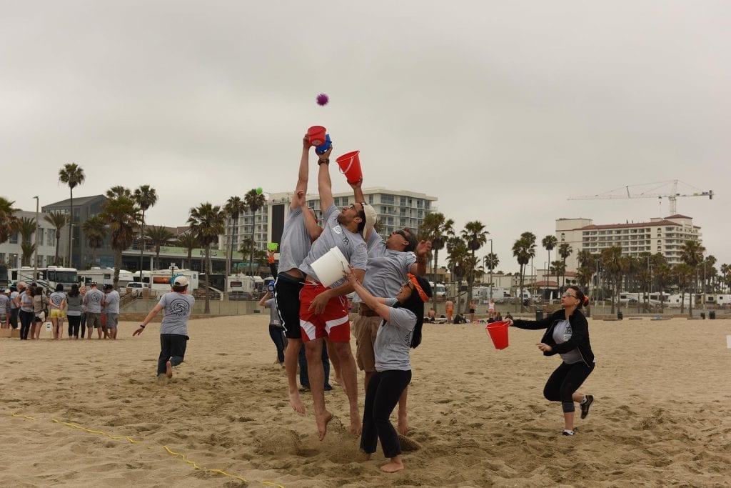 Team members compete in a beach team building game, jumping to catch balls with buckets in a high-energy relay challenge.