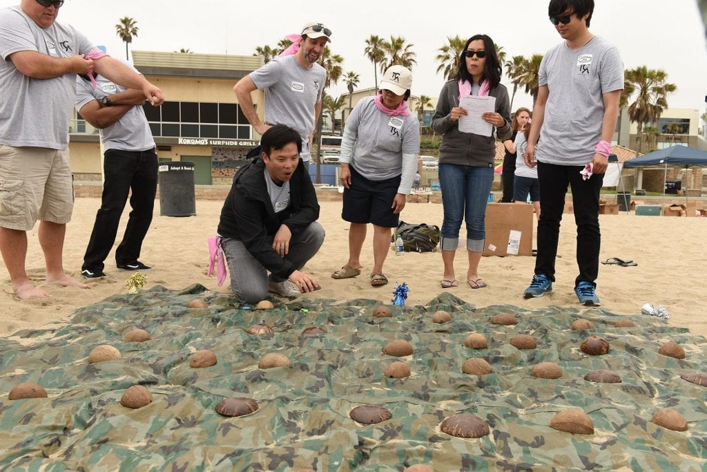 Team members engage in a problem-solving activity on the beach, analyzing and strategizing over a grid of covered items during a team building challenge.