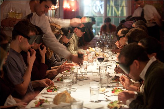 Large group of participants at a 'Dining in the Dark' event seated around a long dining table, wearing blindfolds as they enjoy a meal served by waitstaff, focusing on enhancing their sensory experience through taste and smell.