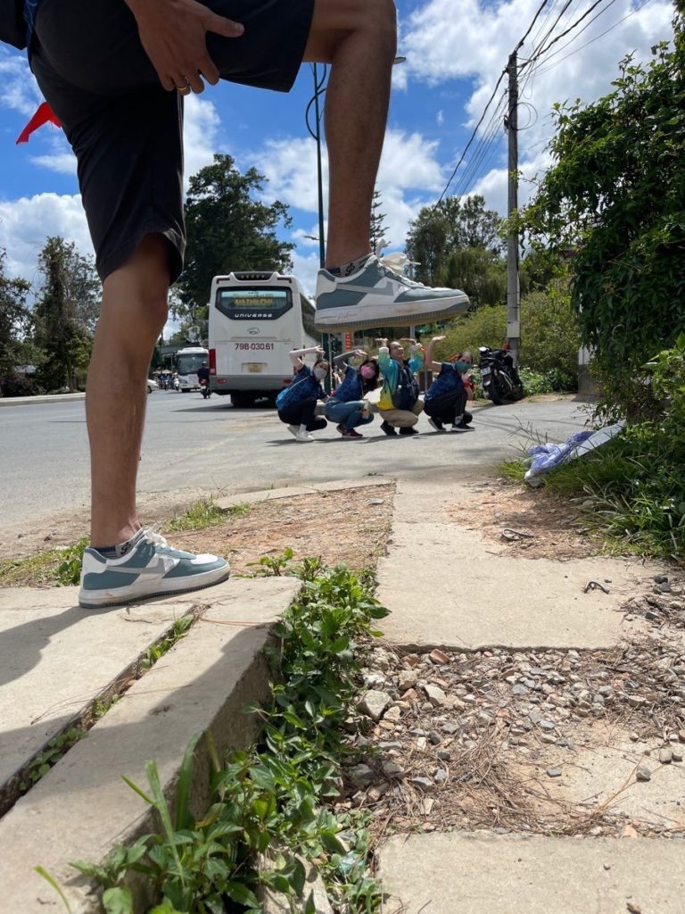 A creative perspective shot of a person lifting their foot, while a group of people crouch in the background during a team building scavenger hunt activity near a roadside.