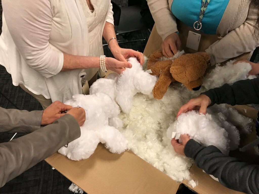 A group of participants are seen from above working together to stuff teddy bears. Their hands are holding various plush bear parts while reaching into a large cardboard box filled with stuffing material. The teddy bears are in different stages of assembly, with some already partially stuffed. The focus is on teamwork and collaboration as they fill the bears for the Team Teddy Rescue Bear team building event.