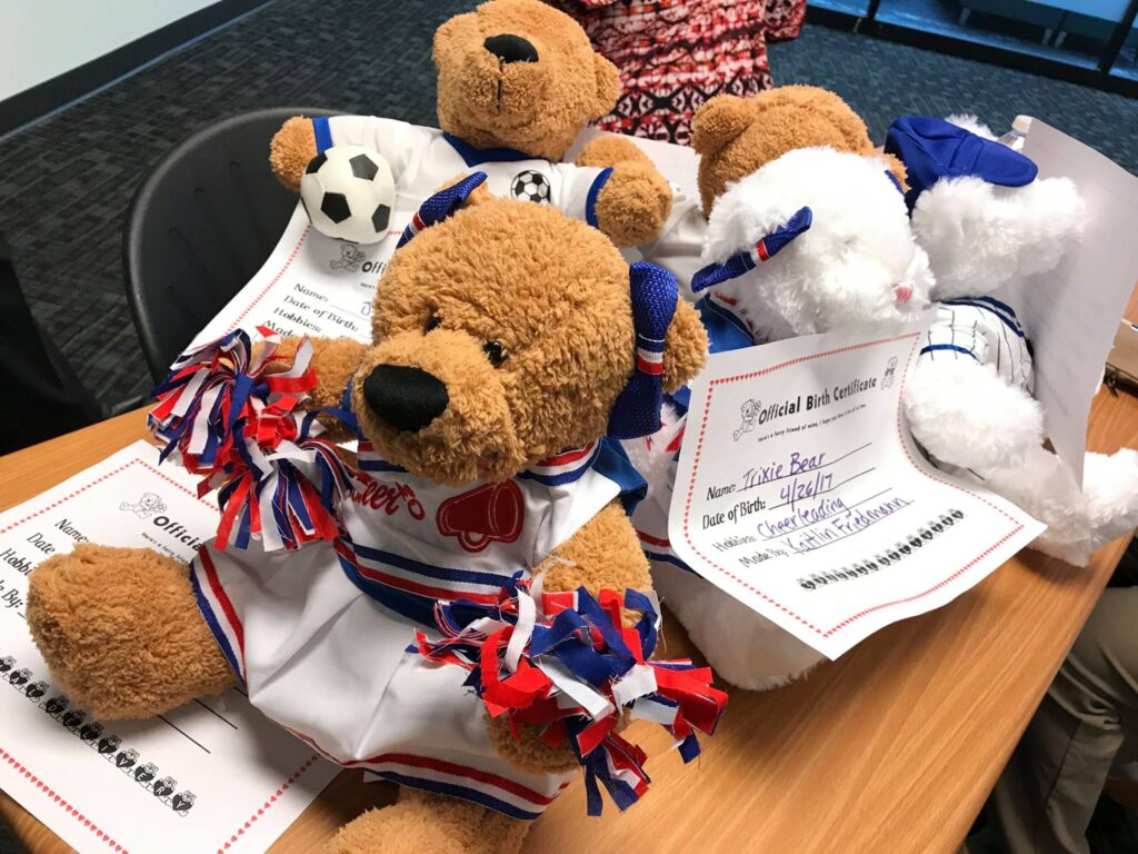 A collection of completed teddy bears dressed in sports-themed outfits, including one as a cheerleader with pom-poms and another as a soccer player with a soccer ball. Each bear has an 'Official Birth Certificate' placed underneath it with handwritten details like the bear's name and date of birth. The scene captures the results of a teddy bear-building activity, part of a charitable team building event.