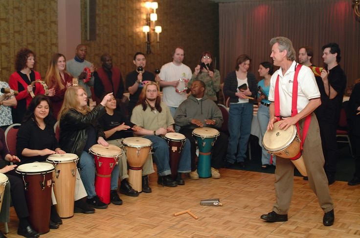 A lively Drumming Up A Team session in progress, with participants actively playing various percussion instruments and a facilitator leading the group in the background.