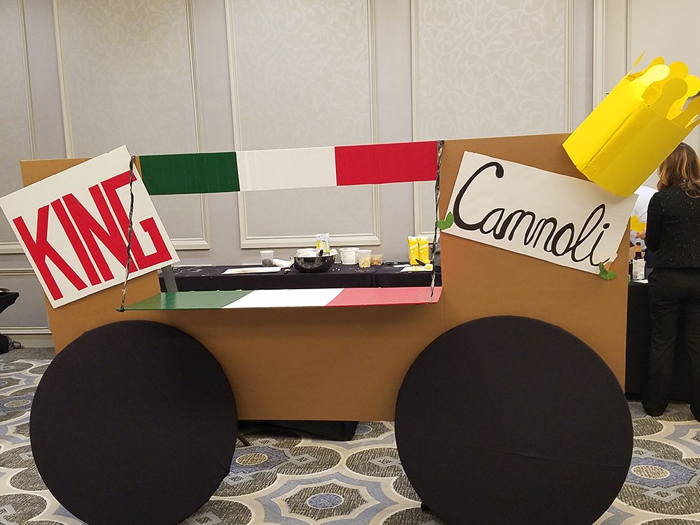 An imaginative 'food truck' display titled 'King Cannoli' is set up as part of a team building event. The mock food truck is constructed with a cardboard facade, and the colors of the Italian flag decorating the upper edge. This creative setup reflects the team's effort to embody an Italian pastry theme for the 'Food Truck Challenge'.