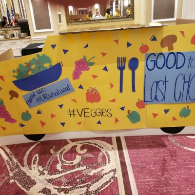 A colorful, handmade food truck façade created for the 'Food Truck Challenge' team building event. The bright yellow design features cut-out images of fruits and vegetables, along with slogans like 'Good to the Last Chop'. The creative display emphasizes a healthy, veggie-focused theme, highlighting the event's blend of culinary skills and artistic expression.