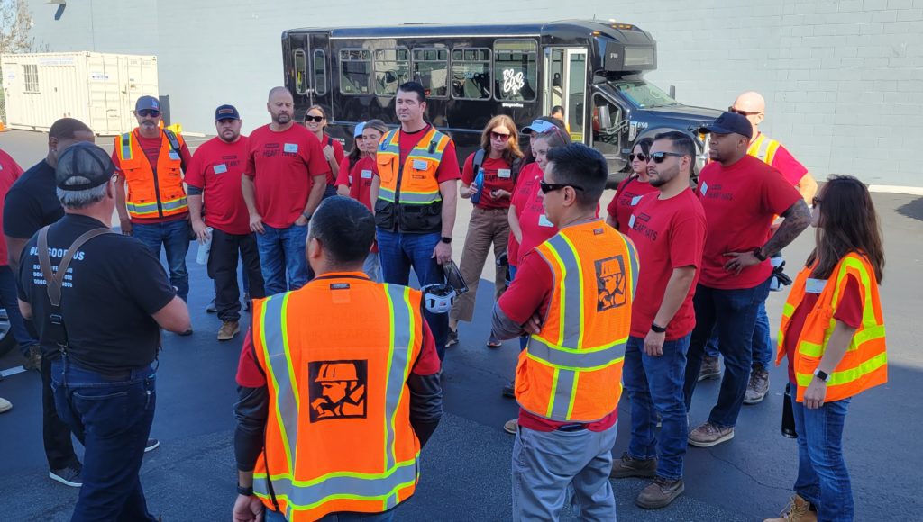 A group of people stands in a circle outdoors, wearing bright orange and yellow reflective safety vests and red shirts. They are receiving instructions before engaging in a charitable team building activity. In the background, a black bus labeled 