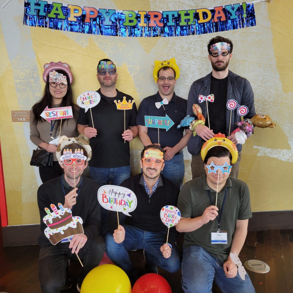 Group of participants posing with fun birthday-themed props and wearing party accessories in front of a 'Happy Birthday' banner during the Birthday Kits activity at a corporate team building event, called 'The Donation Station'.