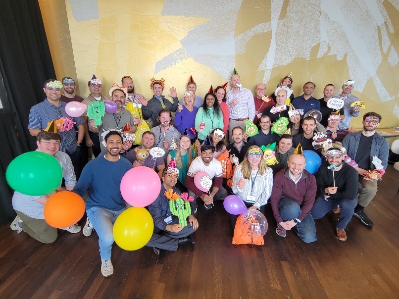 Large group of participants smiling and holding balloons and birthday-themed props, wearing party hats during the Birthday Kits activity at a corporate team building event, called 'The Donation Station'.