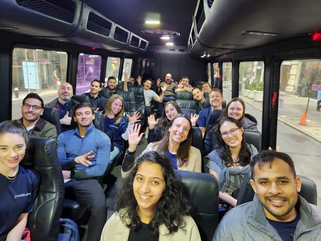 A group of smiling participants sits inside a bus, waving at the camera during a Do Good Bus team building event. The participants are dressed casually, ready to engage in a community service activity.