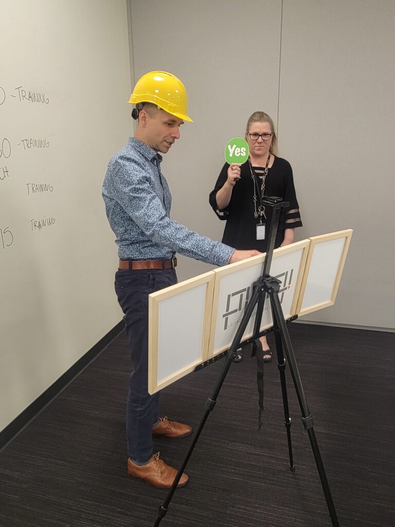 Team building exercise focused on communication and problem-solving. A participant wearing a hard hat reconstructs a model while receiving 'yes' or 'no' feedback, emphasizing the importance of clear instructions and teamwork.