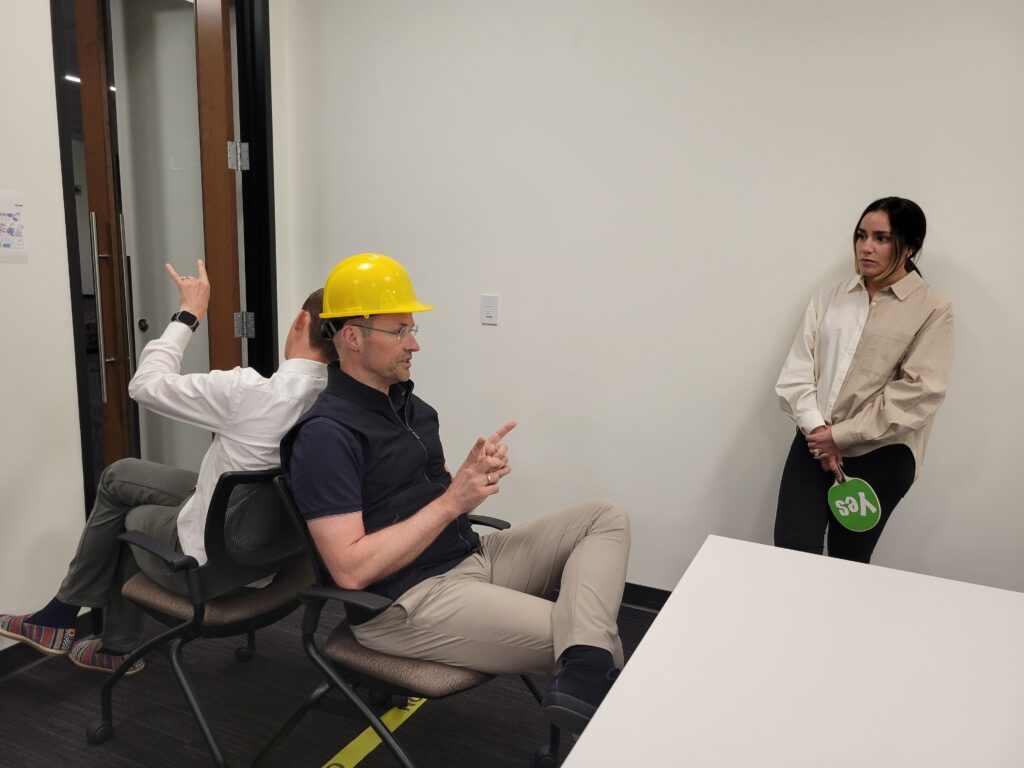 Team building exercise focused on non-verbal communication. A participant wearing a hard hat uses gestures to convey a message while a teammate, seated back-to-back, interprets the instructions, emphasizing teamwork and clear information transfer.