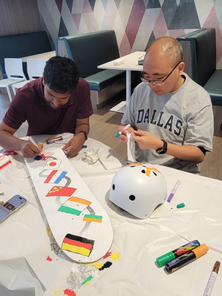 Two individuals at a corporate team building event are focused on decorating a skateboard and a helmet. The skateboard features flags from different countries, while the helmet is being personalized with creative patterns. They are seated at a table covered with art supplies and markers, working collaboratively in a casual environment.