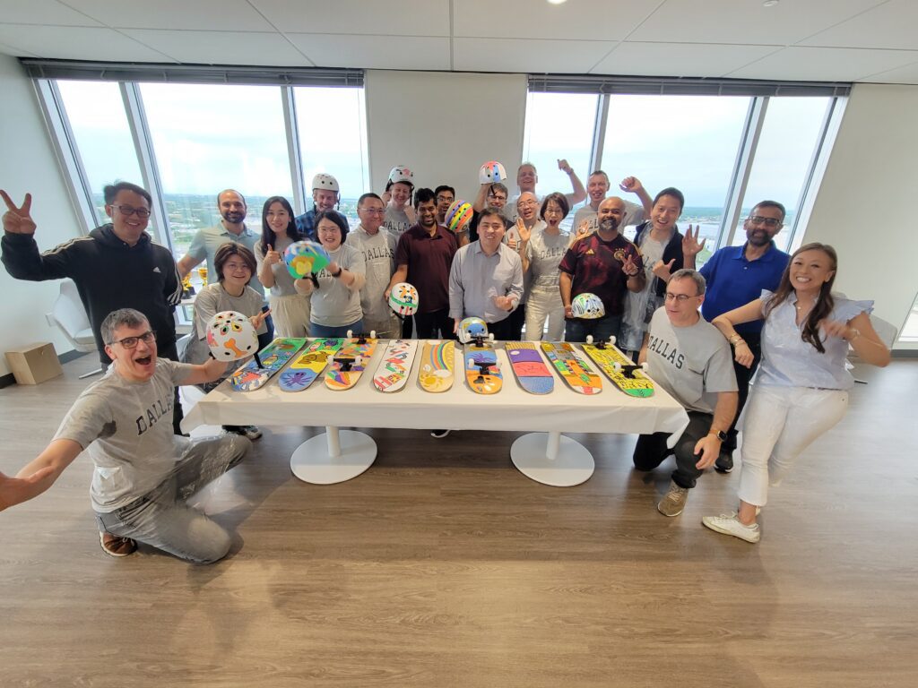 A large group of people celebrating around a table displaying their custom-decorated skateboards and helmets. The skateboards are creatively painted with various colorful designs. Some participants are posing with helmets, and the group looks cheerful, signaling a successful team building event.