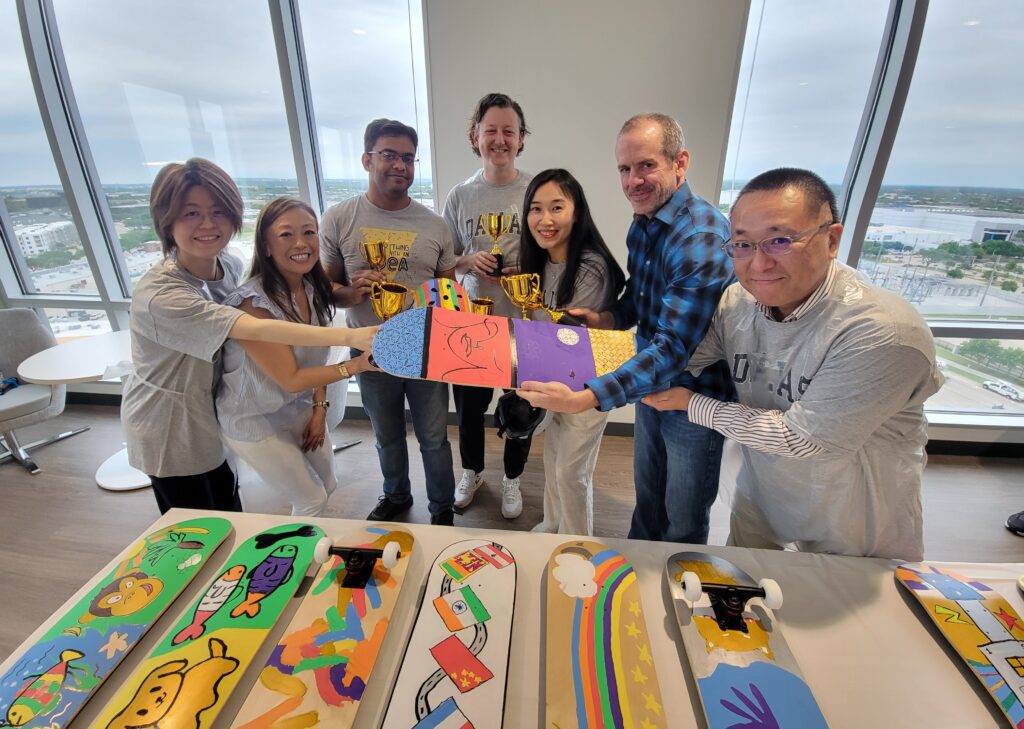 A group of seven individuals smiling and holding up a vibrantly decorated skateboard in a corporate office setting, with large windows. The table in front of them displays several more creatively painted skateboards, each featuring unique designs and bright colors, as part of a charitable team building event.