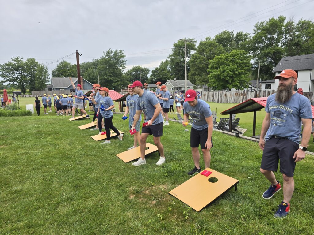 The image shows a group of participants engaged in an outdoor team building activity, specifically playing a game of cornhole. The game involves tossing bean bags into the hole on a raised platform.