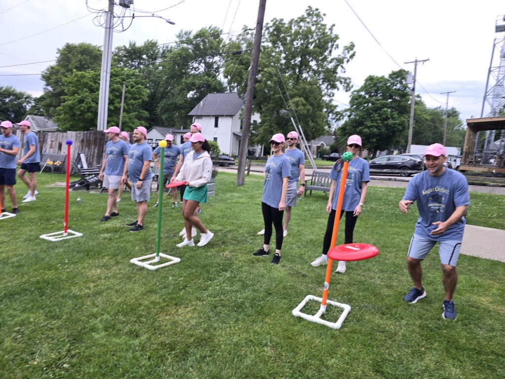 The image shows a group of participants engaged in a team building activity outdoors. The participants are lined up in rows on a grassy field. The activity involves tossing rings or discs onto poles with colored targets. The setup suggests a game that requires accuracy and coordination, promoting teamwork and friendly competition.