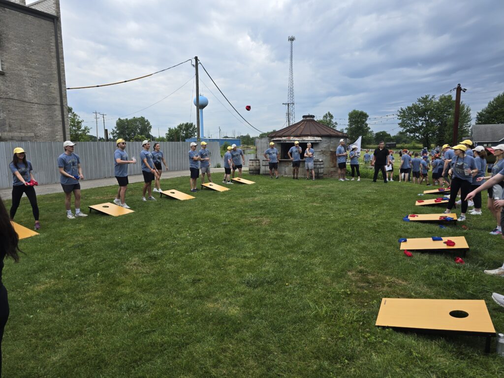 The image shows a group of people participating in an outdoor team building activity. The participants are engaged in a game of cornhole. They are lined up in pairs, tossing bean bags towards wooden boards with holes cut into them.