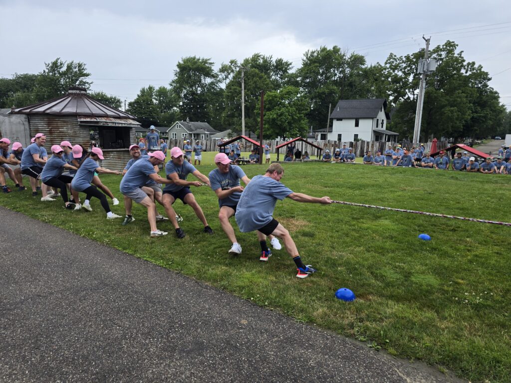 The image shows a group of people participating in a classic game of tug-of-war during an outdoor team building event.