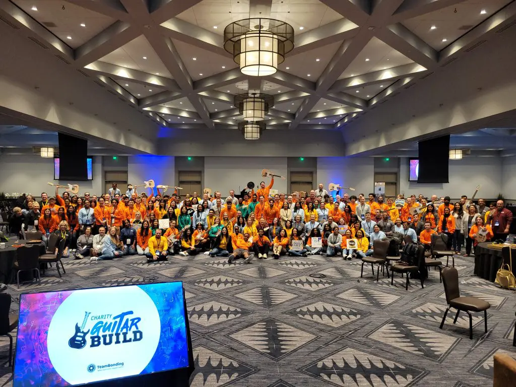 Large group photo at the Charity Guitar Build team building event, with participants holding guitars and celebrating teamwork and community impact.