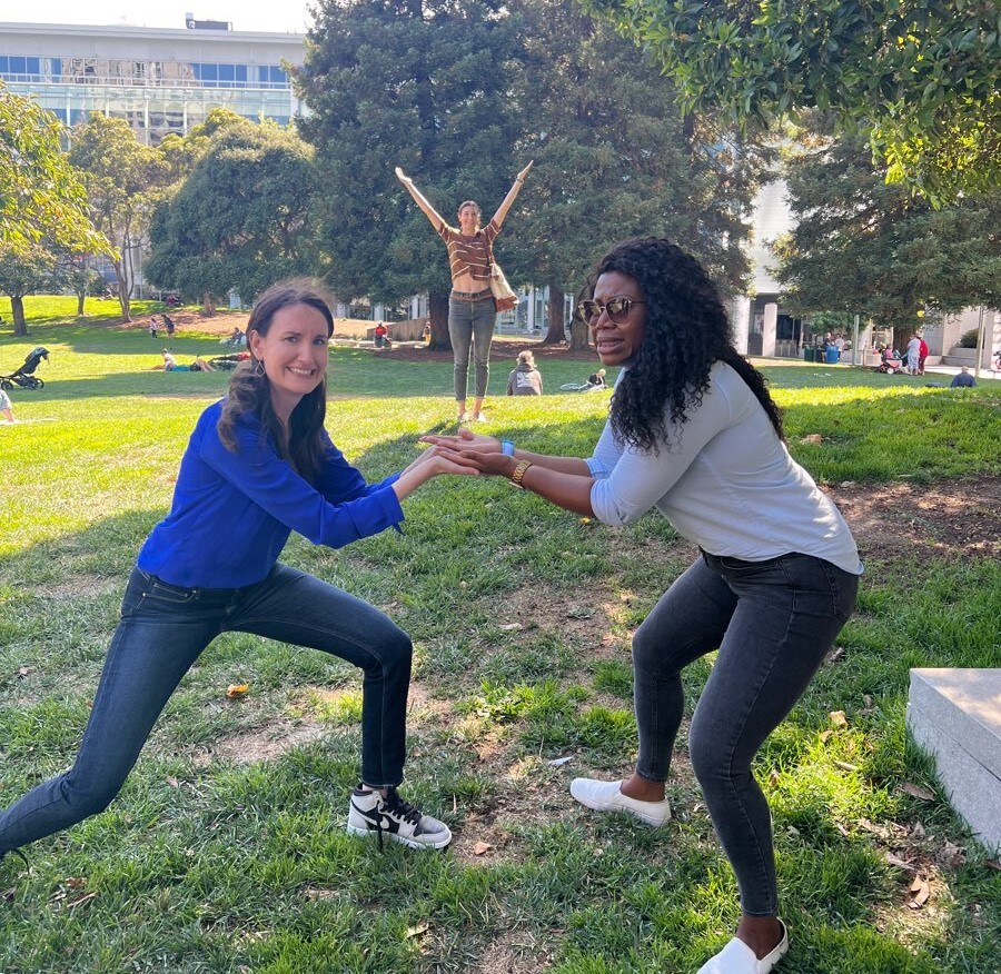 Two women participate in a playful team building activity in a park. They are posing while balancing something in their hands, with a third person standing in the background with arms raised in celebration.