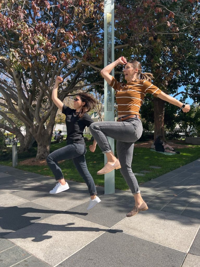 Two women energetically jump in the air during a team building activity outdoors, with trees in the background. Both are striking playful poses mid-jump, adding a sense of fun and excitement to the moment.