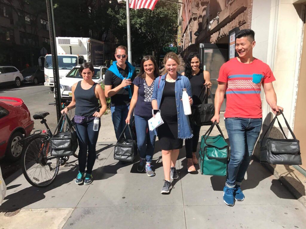 A group of six people walking down a city sidewalk, smiling and carrying insulated bags. They are engaged in a team building activity, possibly delivering meals or supplies.