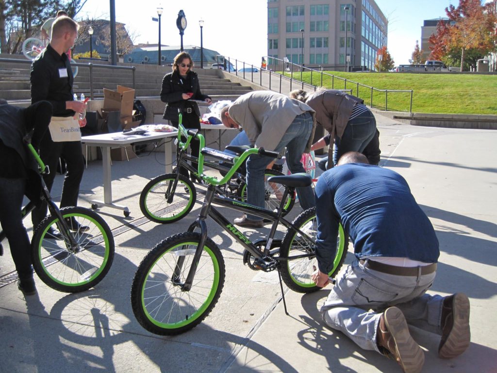 Group of participants in an outdoor Charity Bike Build team building event. Some members are kneeling to work on the bikes, while others stand nearby, discussing instructions. The event is taking place in an open urban space.