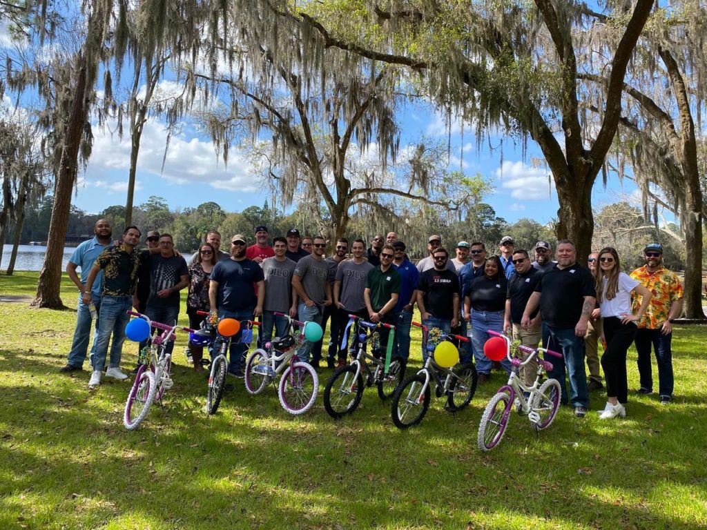 Large group of participants posing outdoors with completed bikes from a Charity Bike Build event. Each assembled bike is decorated with colorful balloons. Participants are smiling in a celebratory mood after a successful team building activity.