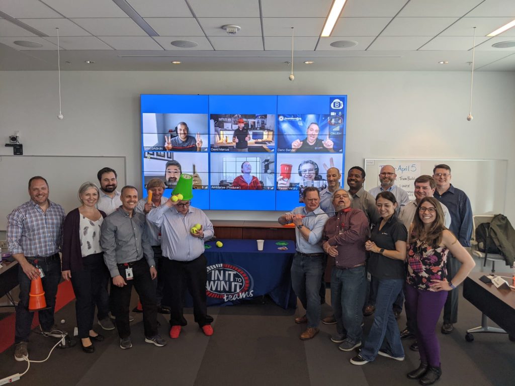 A group of participants posing together with props, smiling and enjoying the In It to Win It hybrid team building activity, with some team members visible on a large screen via video call, showcasing a blend of in-person and virtual participation.