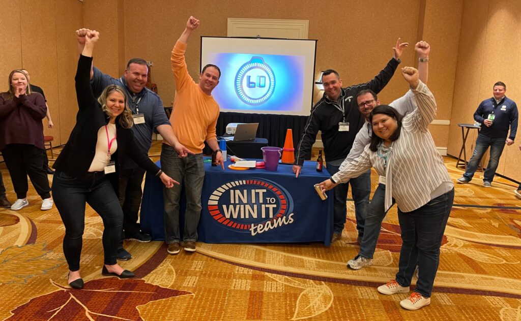 A group of participants celebrating with raised arms and smiling faces in front of a table labeled 'In It to Win It' during a team building activity, showcasing excitement, teamwork, and a fun, competitive atmosphere.