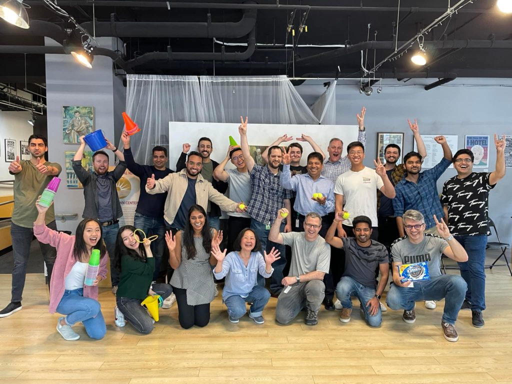 A large group of participants posing energetically with smiles, props, and raised hands, celebrating their participation in the In It to Win It team building event, showcasing a fun and lively group dynamic.