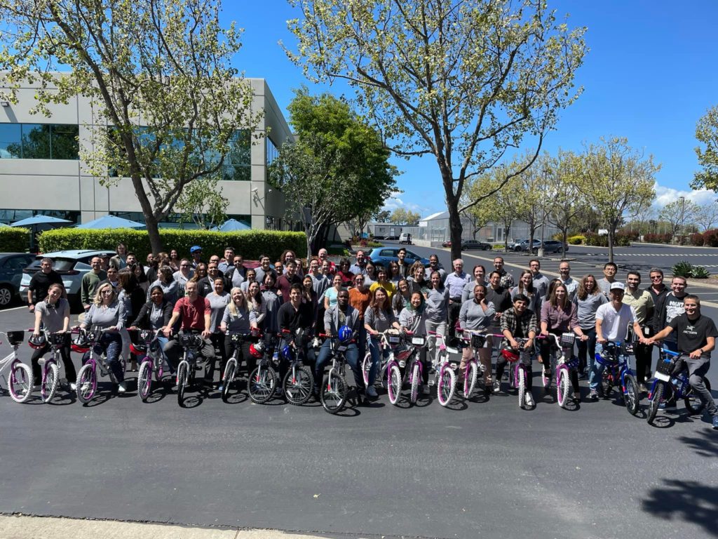 Large group of participants posing outdoors with assembled bikes during a Charity Bike Build event. The group stands proudly behind the rows of bikes, enjoying the sunny day and the sense of accomplishment after working together for a charitable cause.