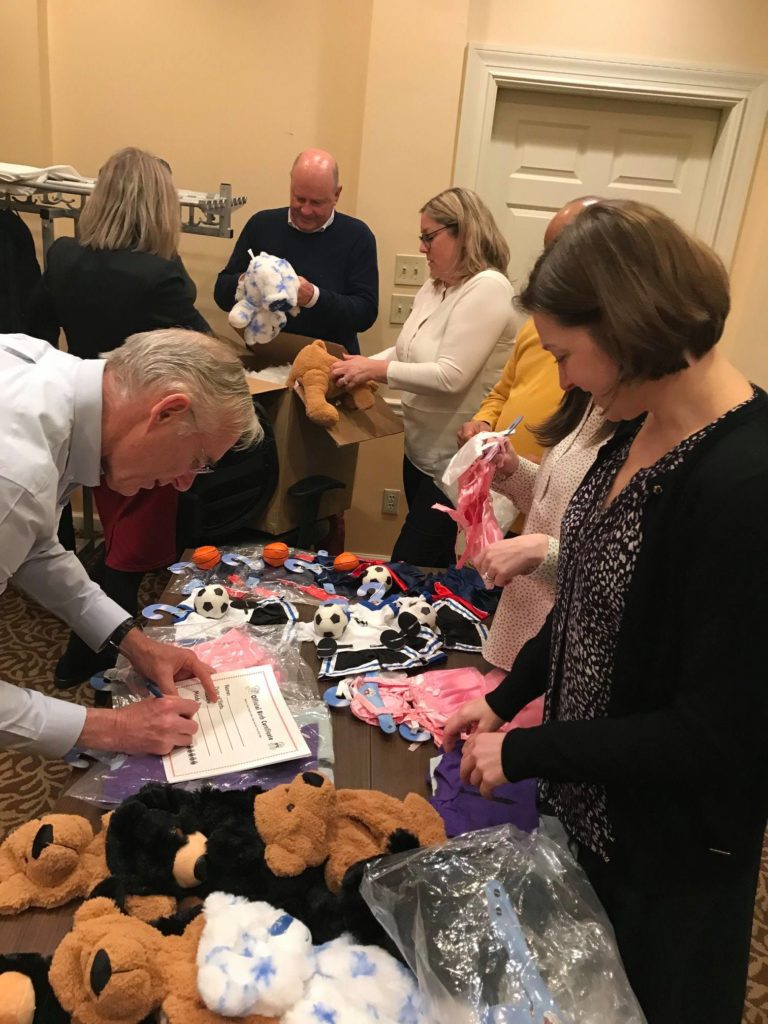 A group of participants actively engage in assembling teddy bears during the Team Teddy Rescue Bear team building event. They are working around a table filled with teddy bear parts, small outfits, and soccer ball accessories. One person is filling out a certificate, while others are selecting outfits and assembling the bears.