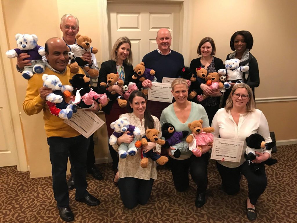 A group of nine smiling participants proudly display the teddy bears they have created during the Team Teddy Rescue Bear team building event. Each person holds one or more stuffed bears in various outfits and colors.