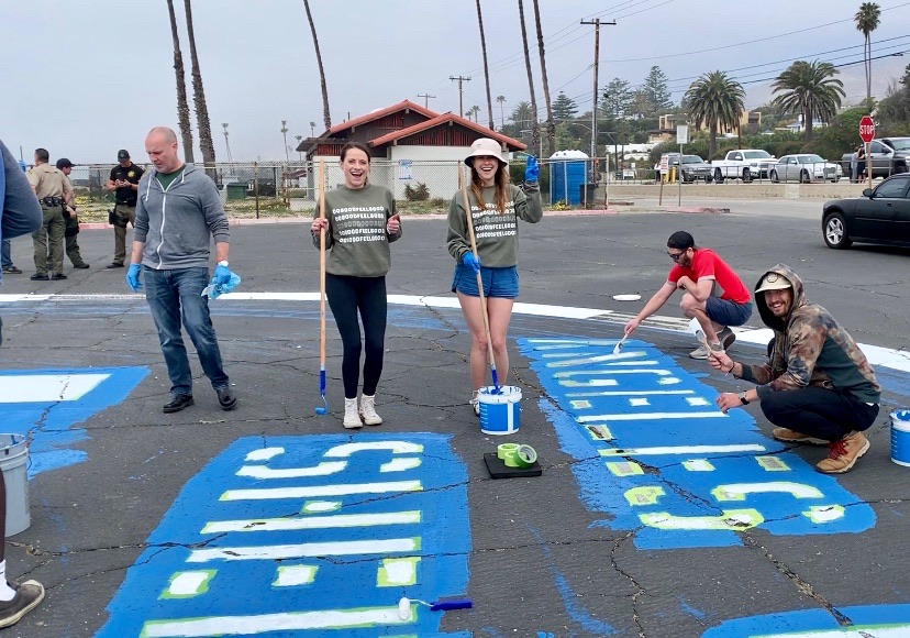 A group of volunteers is painting a large parking lot mural in bold blue and white letters. Two women in the center hold long paint rollers while smiling, and two other volunteers are crouched down applying paint. Palm trees and a beach setting are visible in the background, along with some vehicles parked nearby. Everyone appears to be enjoying the outdoor charitable Do Good Bus team building program.