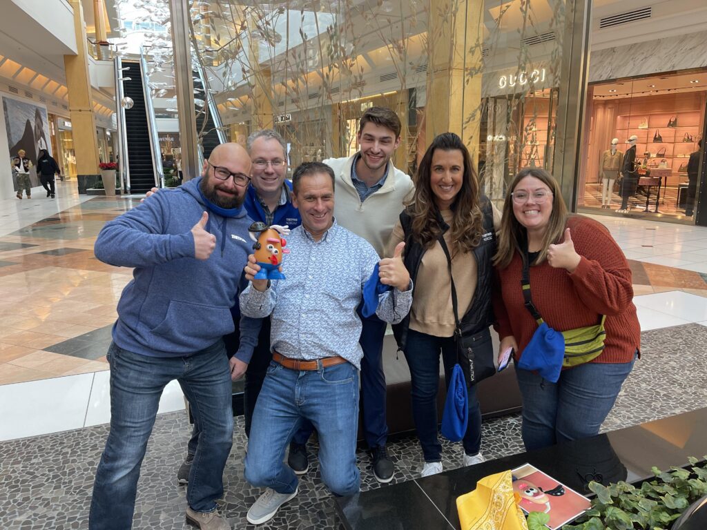 Group of six participants smiling and giving thumbs up during a team building event in a shopping mall, holding a Mr. Potato Head toy.