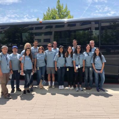 A group of fourteen people standing in front of a large black bus, all wearing matching light blue t-shirts, participating in a team building event. The individuals are smiling and standing close together, indicating camaraderie and a shared purpose.