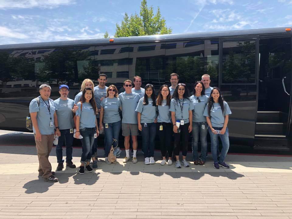 A group of fourteen people standing in front of a large black bus, all wearing matching light blue t-shirts, participating in a team building event. The individuals are smiling and standing close together, indicating camaraderie and a shared purpose.