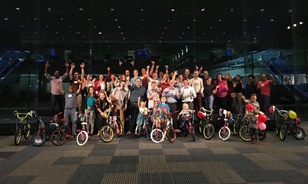 Group of participants smiling and cheering indoors during a Charity Bike Build event. In the front, children excitedly stand with their newly built bikes, surrounded by colorful balloons. The event celebrates teamwork and community engagement as the group successfully completes the charity project.