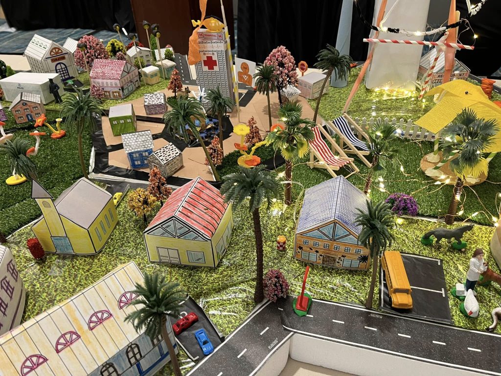 Close-up view of a vibrant City Build team building project showcasing miniature houses, trees, a school bus, and decorative lights on a grassy layout.