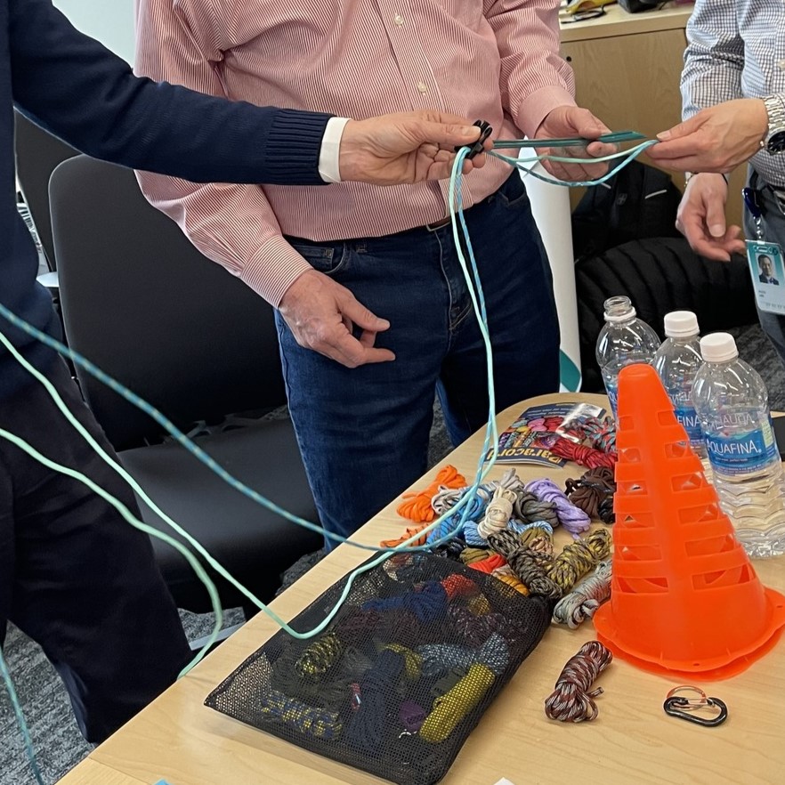 Participants working together with ropes and supplies during the Foster Pet Care Kits activity at a corporate charitable team building event called 'the Donation Station', with various materials and water bottles on the table.