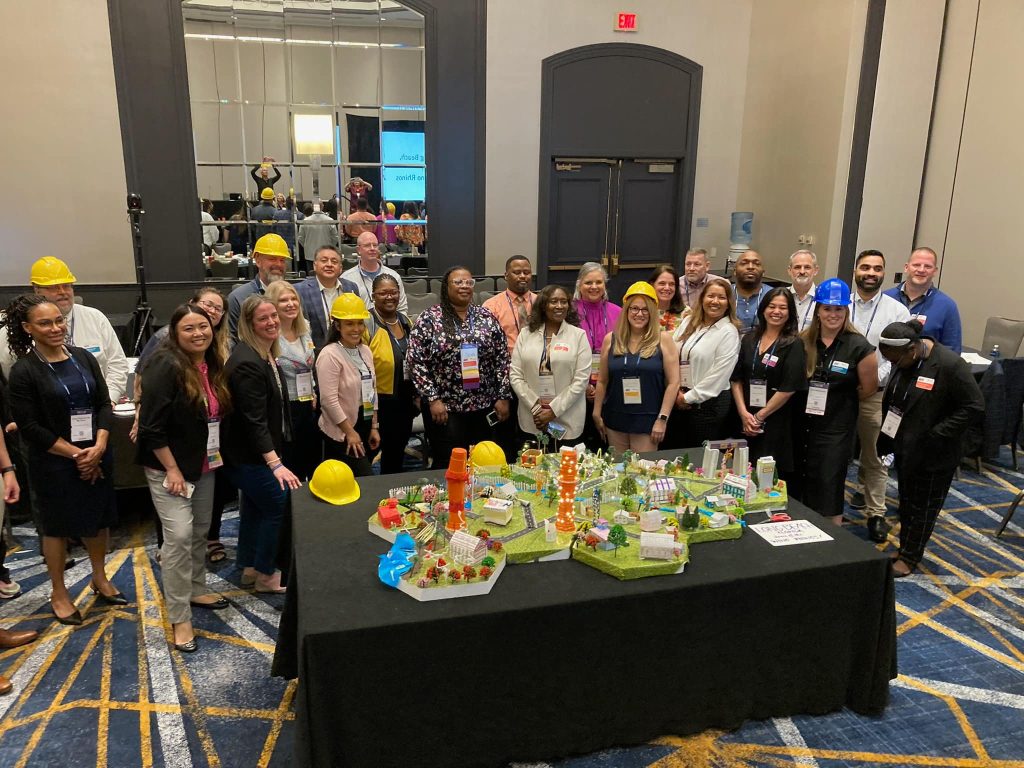 Team members in hard hats pose together around a completed City Build project, showcasing a colorful miniature city model with roads, buildings, and greenery.