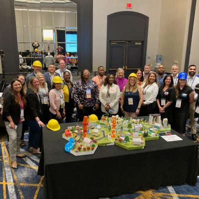 Team members in hard hats pose together around a completed City Build project, showcasing a colorful miniature city model with roads, buildings, and greenery.