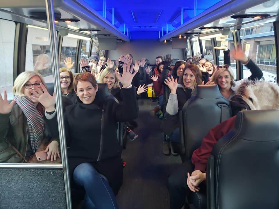 A group of people are sitting inside a bus, smiling and waving enthusiastically towards the camera. The bus is filled with happy, excited individuals, suggesting a fun and engaging group team building event, part of the Do Good Bus program.