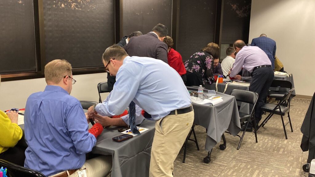 Teams of participants are gathered at tables in a professional setting, focused on assembling prosthetic hands during a team building event. The participants are paired up, wearing red empathy sleeves, and working together on the project. The room has a collaborative atmosphere, with individuals helping each other as they handle tools and components.