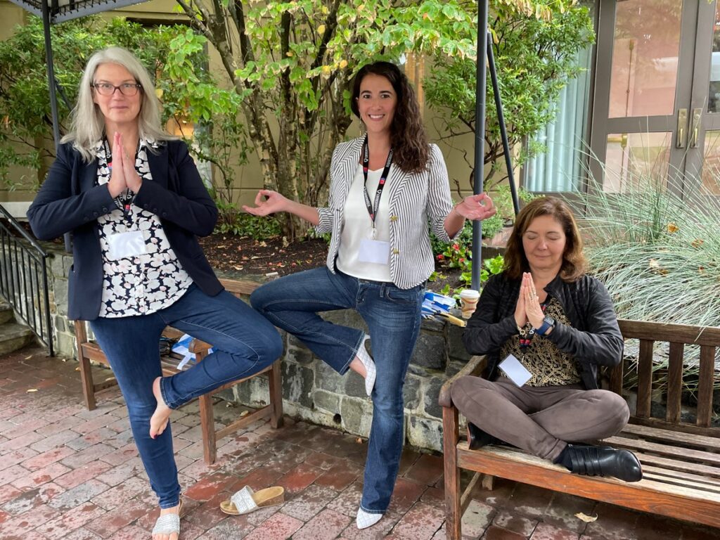 Three team members engage in a relaxing yoga pose outdoors during the Healthcare Heroes team building event. Two participants stand in tree pose while the third sits cross-legged on a bench with her hands in prayer, representing mindfulness and relaxation. This interactive experience embraces Corporate Social Responsibility, helping teams collect items for donation to local hospitals.